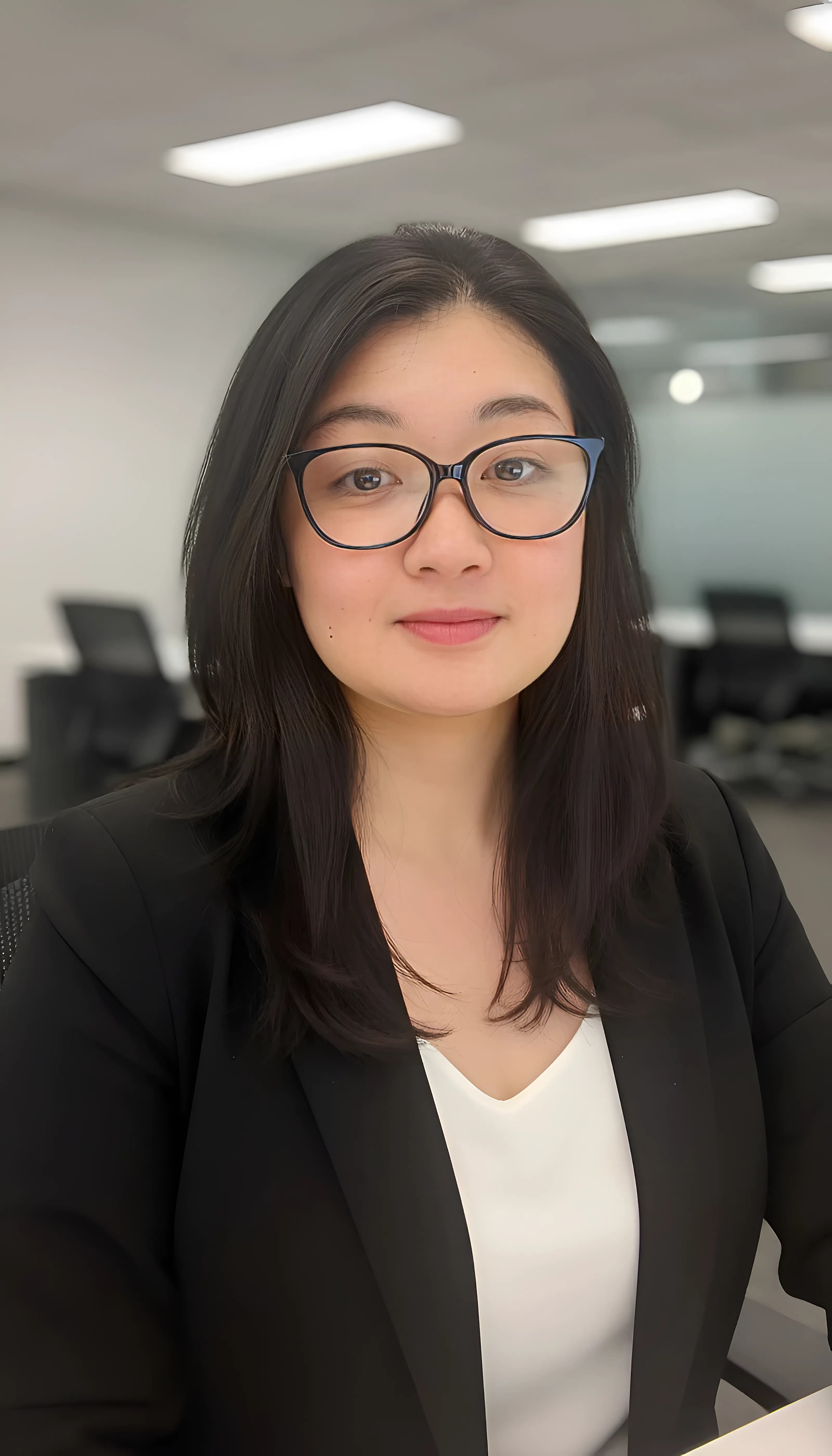 Smiling young woman with dark hair and glasses wearing a black blazer in an office.